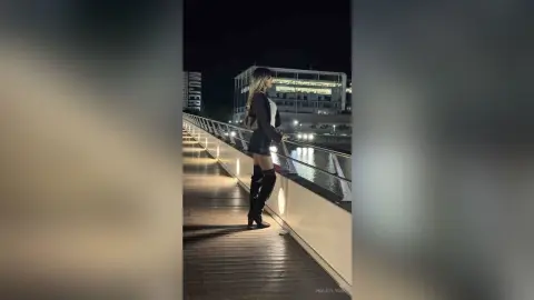Stylish woman in a black mini dress and boots walking on a wooden pier at night near city buildings.