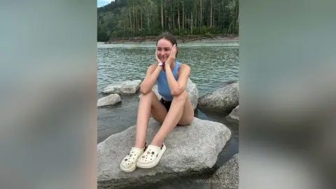 Smiling young woman in a blue tank top sitting on rocks by a peaceful river in a lush forest setting.