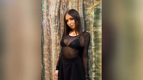 Young woman in a black sheer long-sleeve dress posing against a textured marble wall background.