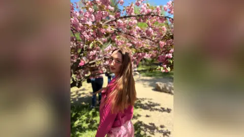 Young woman in a pink jacket smiling under blooming pink cherry blossom trees in a sunny park.