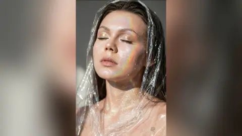 DemetriceTaneja poses with a wet, shiny look under a clear plastic wrap, showing her face and shoulders with a rainbow light reflection.