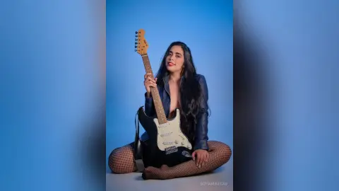 Young woman in a blue leather jacket and fishnet tights holding an electric guitar against a blue background.