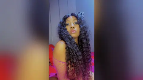 JoanJon with long curly hair wearing a pink fishnet top while posing for the camera.