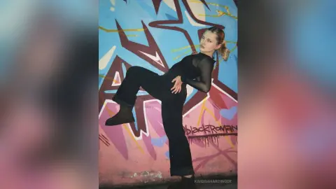 Young woman in black mesh top and dark jeans posing confidently against a colorful graffiti wall.