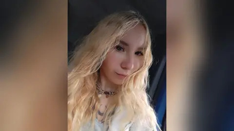 Blonde woman with wavy hair wearing a silver necklace in a soft, natural light portrait.