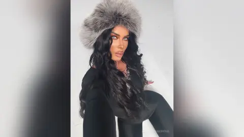 Glamorous woman with long dark hair wearing a grey faux fur hat and black outfit in a bright studio setting.