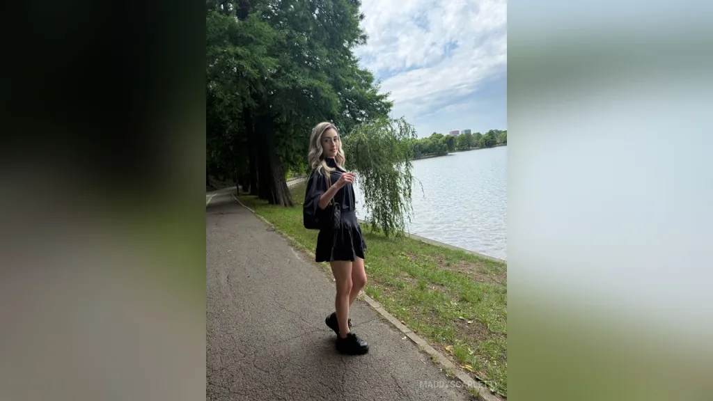 Maddy — MaddyScarlett stands on a park path wearing a short black dress and black shoes with trees and a lake in the background.