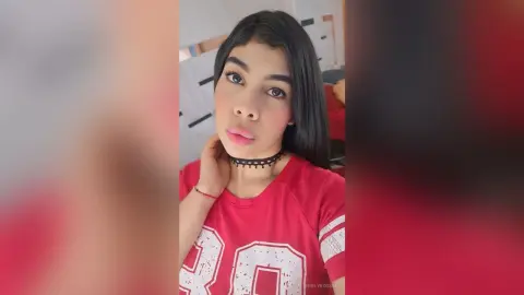 Young woman with long dark hair wearing a red graphic t-shirt and black choker in a bright indoor setting.
