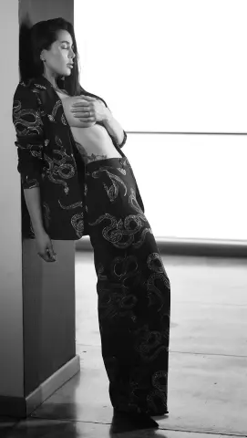 Moody black and white portrait of a woman in a patterned snake print suit leaning against a wall.