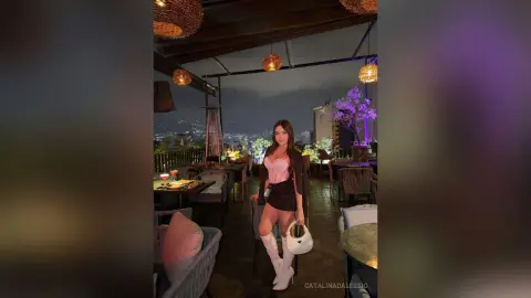 Stylish woman in a pink top and black skirt posing at a rooftop lounge at night under warm ambient lighting.