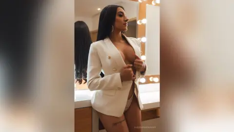 AlessandraSanit poses in a white blazer and lace panties, showing off her cleavage and long black hair in a lit dressing room.