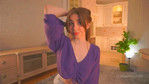 Young woman in a purple ribbed top posing in a cozy, warm-lit modern kitchen setting.