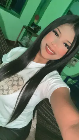 Smiling young woman with long black hair wearing a white graphic t-shirt in a bright green room.