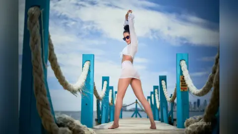 JessieLiu stands on a wooden pier wearing a white crop top and white panties, posing with her arms raised against a blue sky.