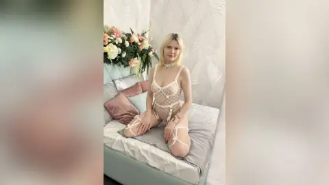 Blonde woman in white lace lingerie and stockings sitting on a bed in a bright, floral bedroom setting.