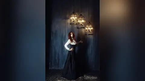 RubyAttwood poses in a long black skirt and white long-sleeved top against a dark blue wall under hanging cage lights.