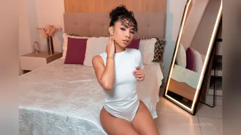 Young woman in a white bodysuit sitting on a bed in a bright, modern bedroom.