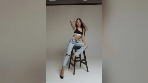 MilanaVolkova poses sitting on a black stool wearing a black crop top and ripped light blue jeans, running her hand through her long brown hair.