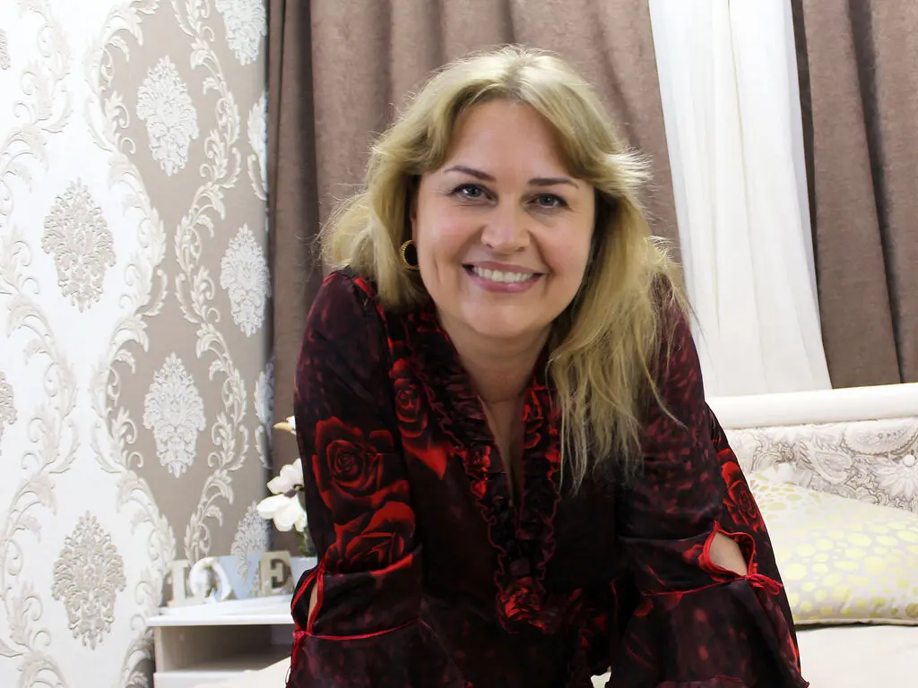 AlanaSharm smiles while wearing a red and black rose-patterned blouse with ruffled sleeves.