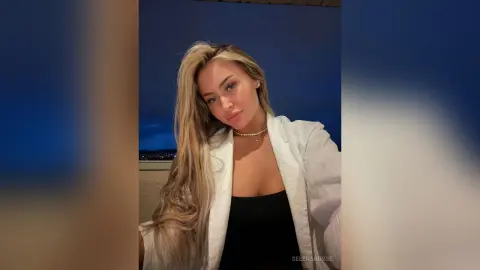 SelenaRusse poses outdoors at night wearing a white blazer over a black top, showing her long blonde hair and cleavage.