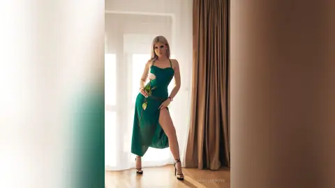 Elegant blonde woman in a sleek emerald green satin dress holding a rose in a bright, minimalist room.