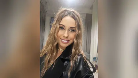 Smiling blonde woman in a black leather jacket posing in a bright indoor setting.