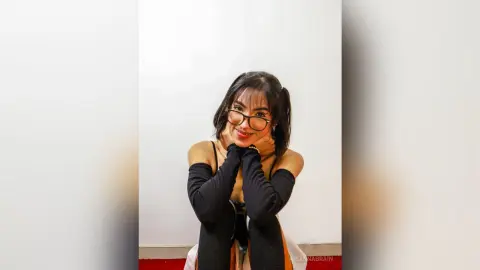 Smiling young woman with glasses in a black sleeveless top and long gloves posing against a white wall.