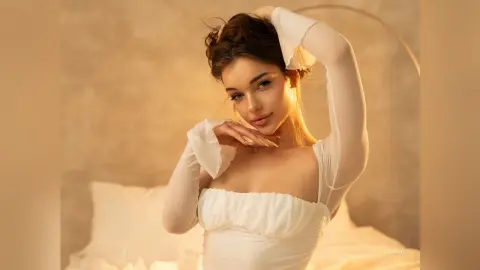 Beautiful woman in a white long-sleeve corset top posing in a warm, cozy bedroom setting.