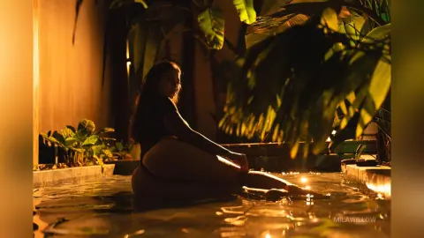 MilaWillow sits naked in a dark tropical pool at night, illuminated by warm golden lights.