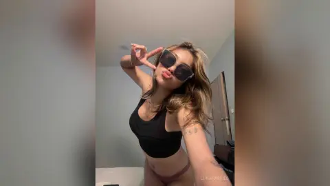 Stylish young woman in a black sports bra and sunglasses making a peace sign in a modern bedroom.