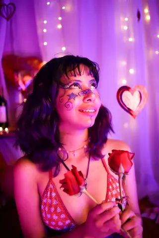 Smiling woman with face art holding red roses in dreamy pink lighting for a romantic Valentine's Day mood.