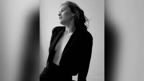 EmilyGiffin poses in a black blazer with nothing underneath, showing her bare chest and neck against a plain light background.