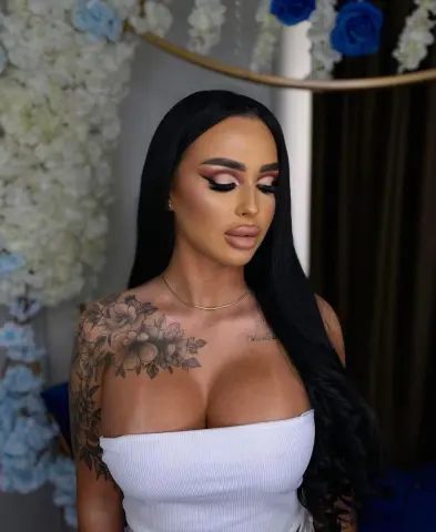Glamorous woman with long black hair and floral tattoos wearing a white tube top in a floral setting.
