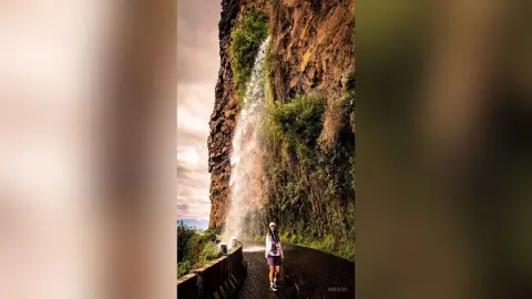 Adventurous female hiker in casual gear standing before a massive tropical waterfall and rocky cliffside.
