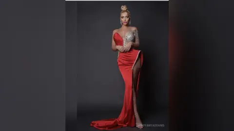 TiffanyAddams poses in a long, red, off-the-shoulder gown with silver detailing and a high slit, standing against a dark grey background.