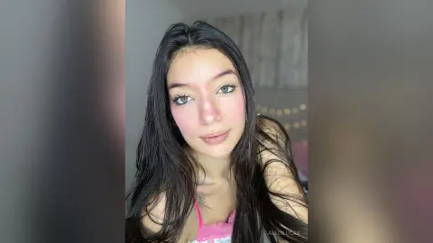 AleDilucas looks at the camera with long dark hair and pink blush, wearing a pink top in a close-up shot.