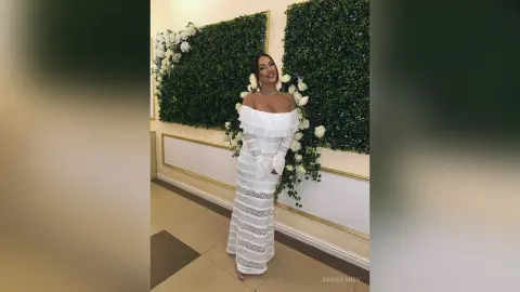 AnnaKarev poses in a long white off-the-shoulder dress standing in front of a green floral wall.