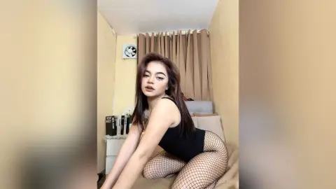 Young woman in a black bodysuit and fishnet tights posing in a cozy bedroom setting.