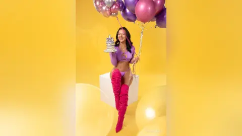 Cheerful woman in a purple crop top and pink boots holding a cake amidst floating balloons on a yellow background.