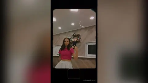 Young woman in a pink crop top and white skirt taking a mirror selfie in a modern room.