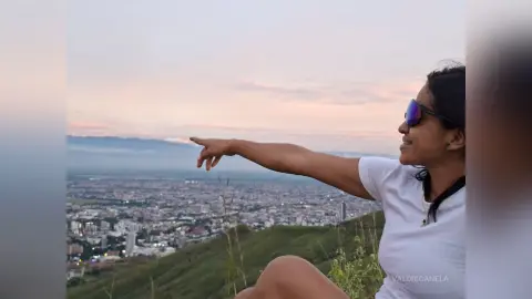 Smiling woman in a white t-shirt pointing at a city skyline during a peaceful sunset on a grassy hill.