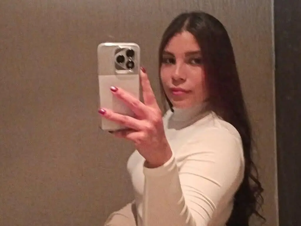 Young woman with long dark hair wearing a white turtleneck taking a mirror selfie in a dimly lit room.