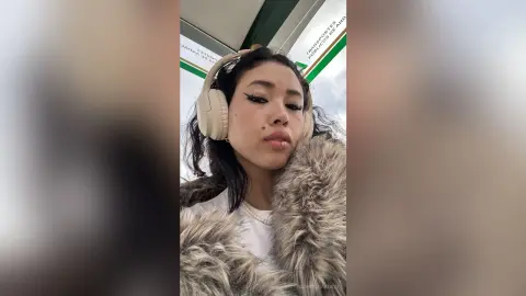 Young woman with headphones and a fuzzy brown coat posing outdoors under a cloudy sky.