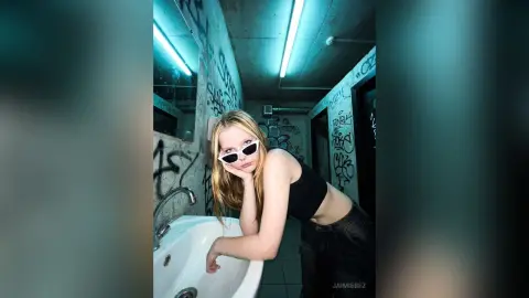 JaimieBez leans over a white sink wearing white sunglasses and a black crop top.