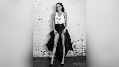 Edgy woman in a white graphic tank top and black flowing skirt posing against a white brick wall.