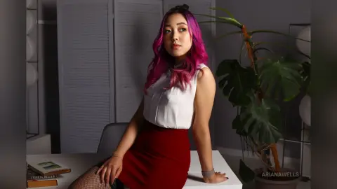 Young woman with pink hair in a white sleeveless top and red skirt sitting in a moody, modern room.