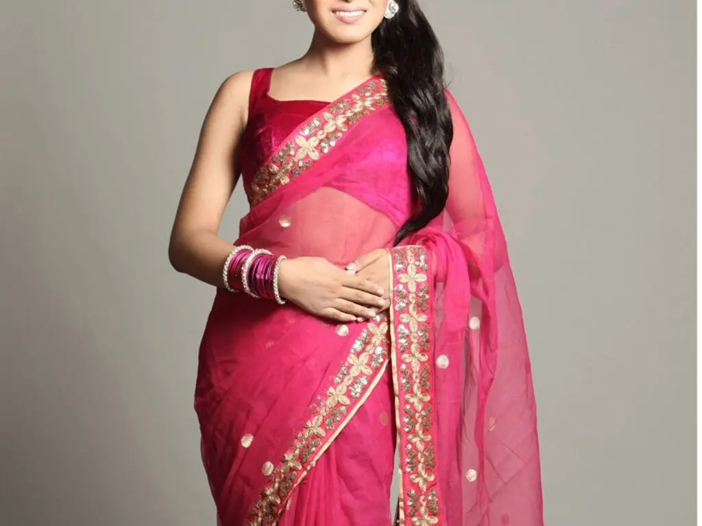 Elegant woman in a bright pink traditional Indian saree with gold embroidery posing against a grey background.