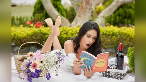 LucyMartin lies on a white blanket in a garden, wearing a white top while reading a red book.