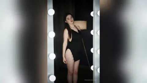 Young woman in a black cutout dress posing in a studio with vanity lights and a moody atmosphere.