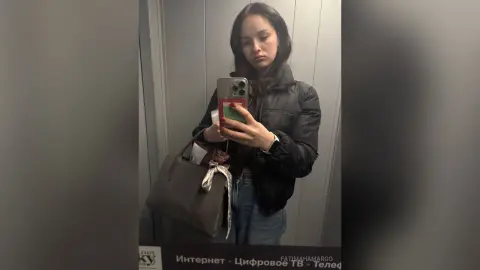 Young woman in a black puffer jacket and jeans taking a moody mirror selfie with a large tote bag.